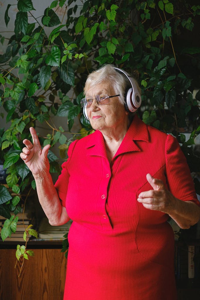 Elderly cheerful female in bright red dress and headphones listening to music while dancing and looking away near green plant