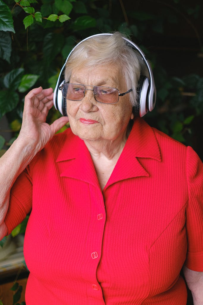 Elderly dreamy female touching wireless headphones while listening to music and looking away in garden