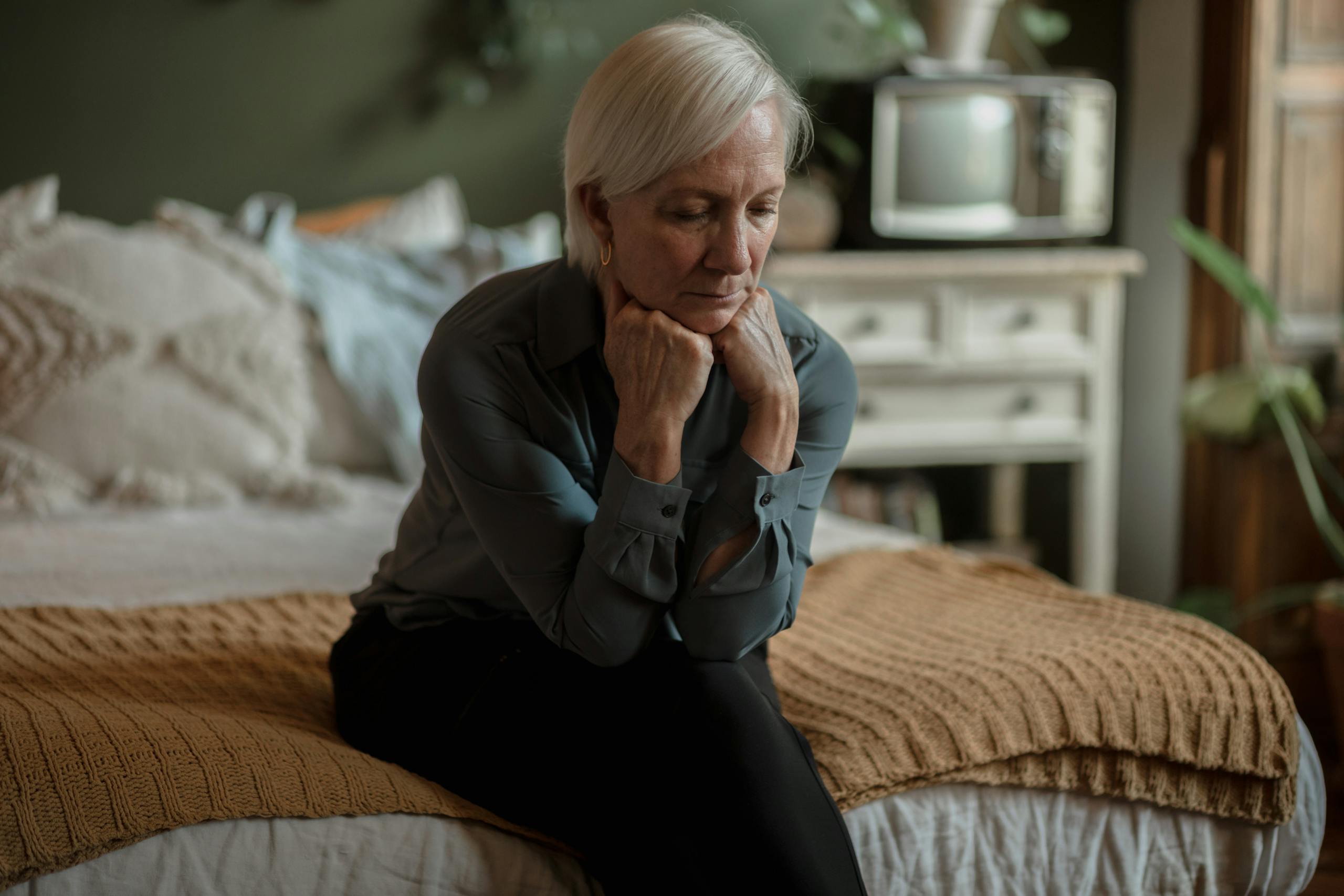 An elderly woman sitting on a bed, deep in thought, with a somber expression in a cozy bedroom.