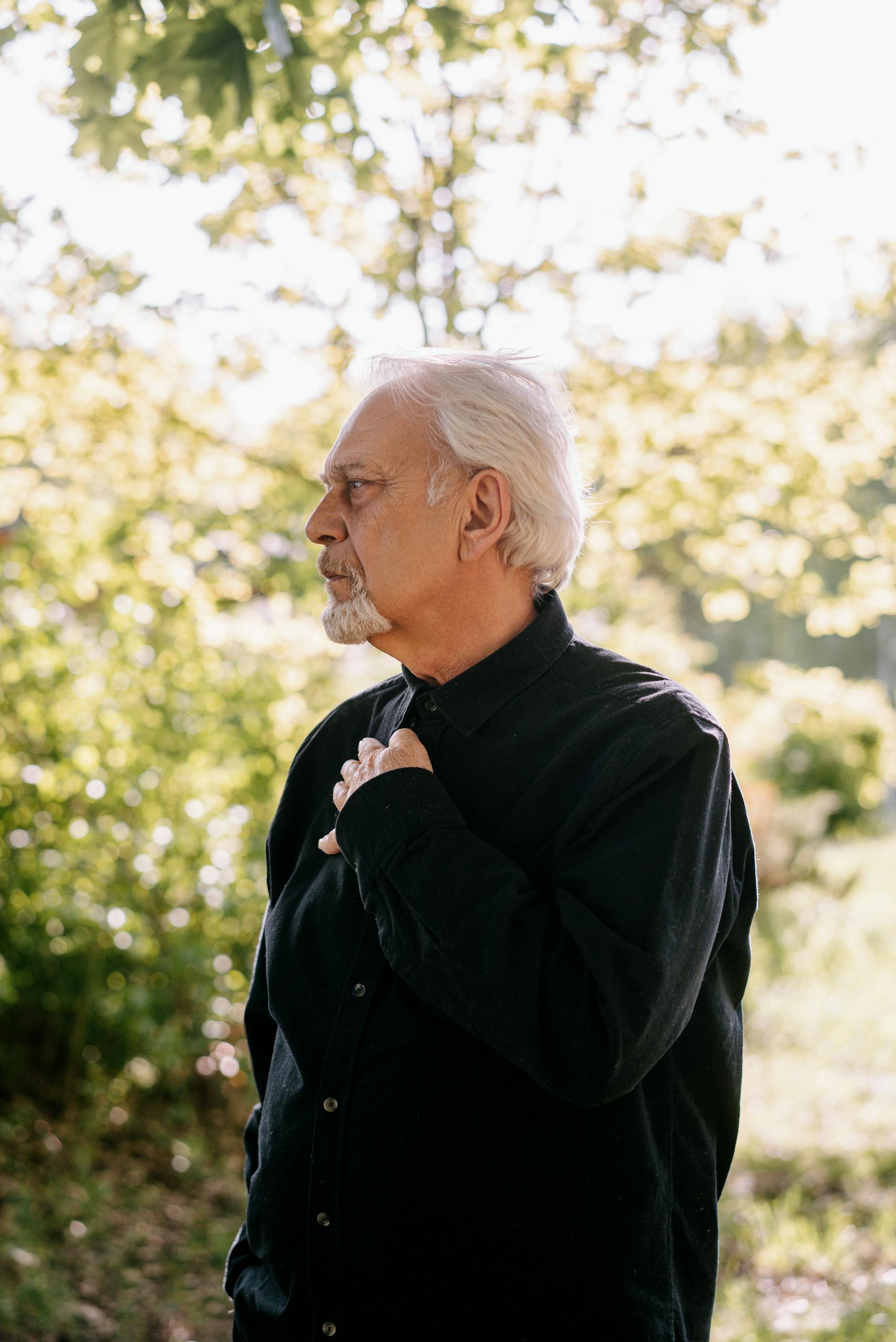 Pensive elderly man with white hair in nature, wearing black clothes, looking contemplative against a sunlit background.