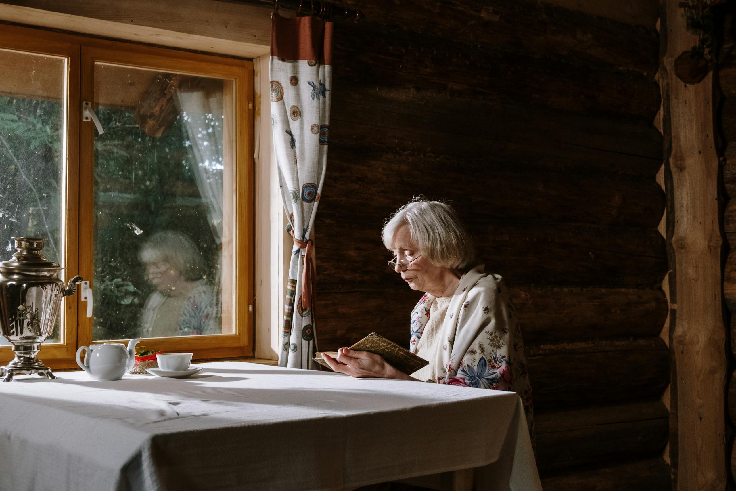 Elderly woman reading a book inside a rustic cabin, enjoying a peaceful moment.
